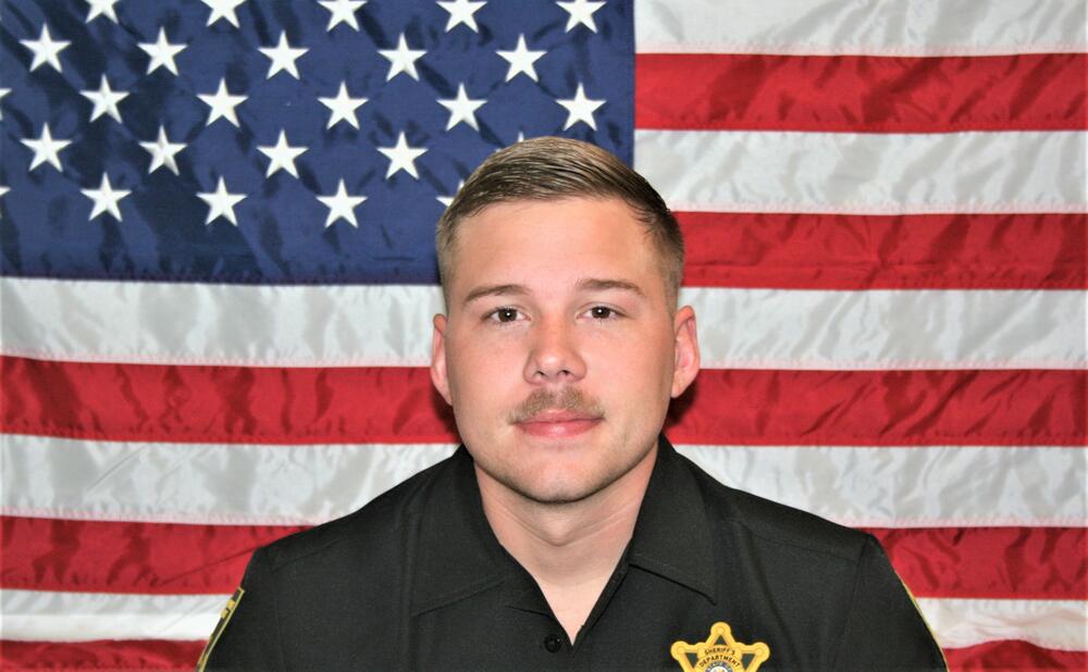 White male with blonde hair and mustache wearing a black polo with a gold Sheriff's badge sitting in front of the American Flag.