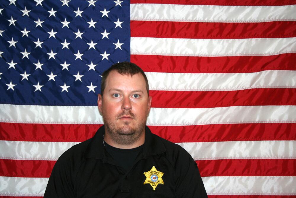 White male with facial hair, black polo with gold sheriff's badge. Sitting in front of the American Flag