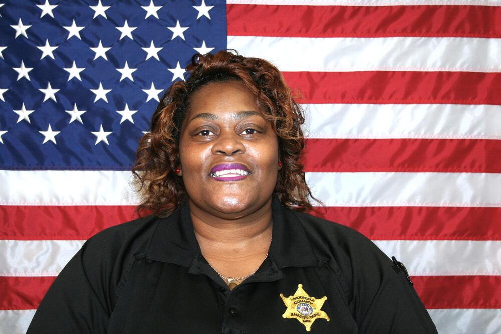 Black female wearing a black polo with a gold sheriff's badge sitting in front of the American Flag.